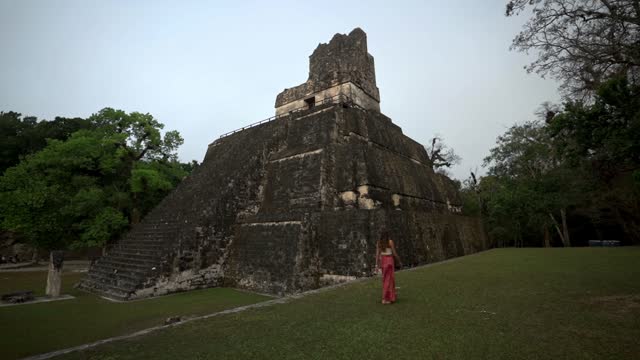 A woman tourist walking across grass observing an ancient temple ruin at Tikal National Park, Guatemala. Slow Motion.