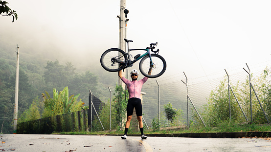 Cyclist lifting the bike over the top of the mountain with his hands celebrating his arrival at the top of the mountain.