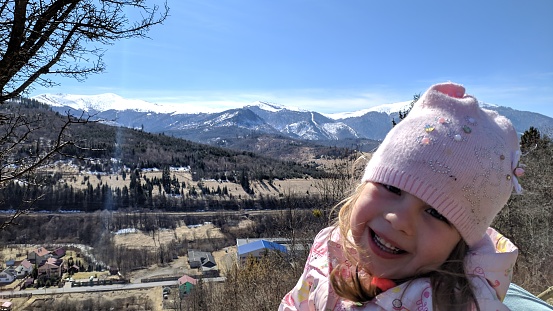 A laughing girl 4-5 years old against the backdrop of house roofs and snow-capped mountains.