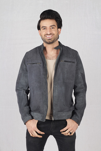 A young male model posing in style with grayish black leather jacket with black top and jeans in white background