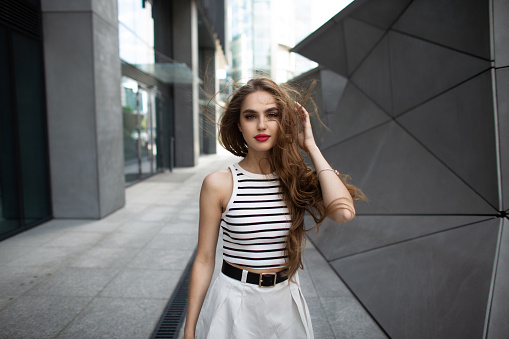 Beautiful young stylish girl model with red lips in fashion summer casual clothes with a top and a skirt walks in the modern city
