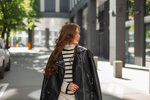 Happy beautiful fashionable woman with a smile in stylish casual clothes with a leather jacket walks on a sunny day in the city