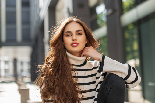 Beautiful European girl model in fashionable clothes with a striped sweater sits in the city on sunny day