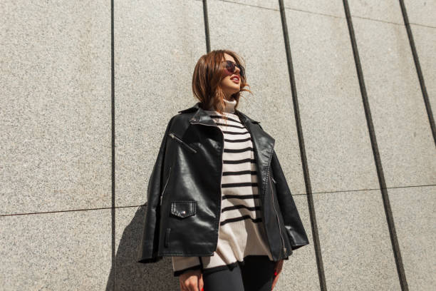 happy beautiful young fashion urban rock girl with sunglasses in a black leather jacket and a stylish striped sweater on a sunny day by the wall - wall women leather street ストックフォトと画像