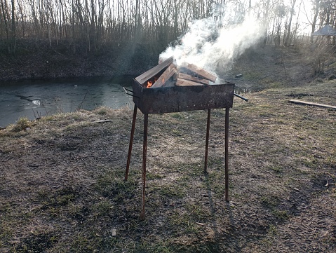 A metal grill on the background of a small pond. A bonfire lit in a brazier made of ash wood from which white smoke rises. Relaxation in nature with cooking on fire.