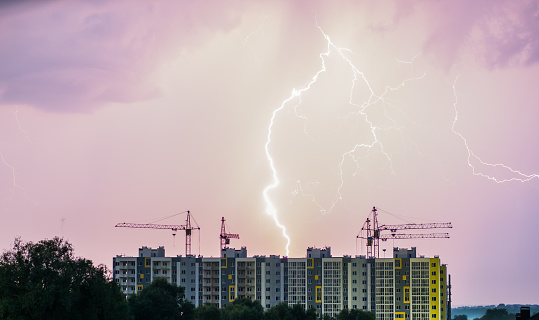 An apartment building with construction cranes against the background of the night sky where you can see lightning.