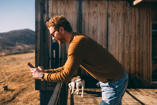 Young adult male texting on phone outside wooden cabin.