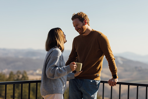 Happy couple sharing a laugh over mugs of coffee with a scenic mountain view in the background, enjoying the moment together.