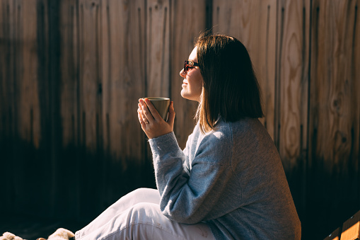 Side view of a calm woman sipping coffee, basking in warm sunlight with a wooden backdrop, emanating tranquility and comfort.