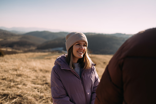 Happy young woman in a winter beanie and jacket smiles while standing in a mountainous landscape, embodying joy and wellness.