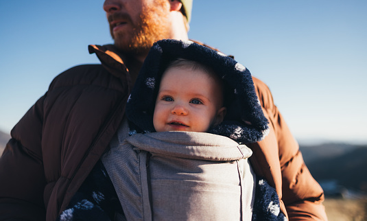 Close-up of a cheerful baby bundled in a carrier during a sunny hike with father against a scenic backdrop.
