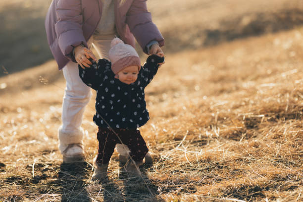 A toddler taking first steps outdoors with the support of an adult on a sunny autumn day, symbolizing growth and care.