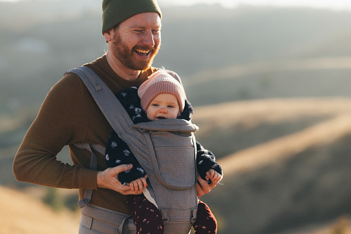 A joyful dad with a beard smiling as he carries his infant in a baby carrier during a hike on a sunny day.