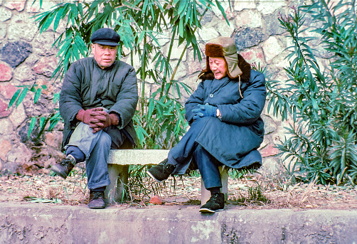 Beijing, China - December 30, 1986: two old men in thick clothes in winter sitting on a bench in front of the wall of forbidden city in Beijing.