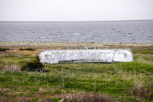 Overturned fishing boat on a grassy meadow near the shore near Cape Ray, Newfoundland with calm sea and grey sky in the background
