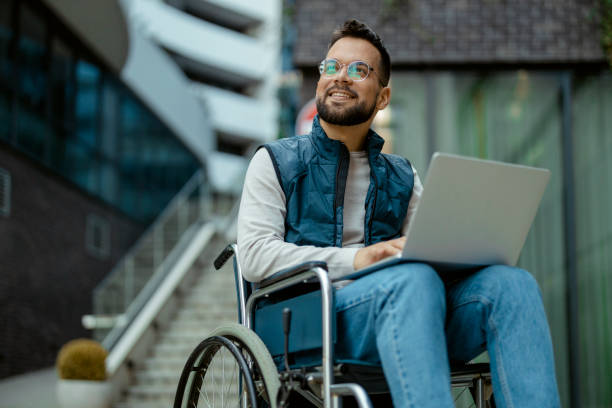 Handicapped Man With Laptop in Wheelchair Outdoors Young Handicapped Man Using Laptop in Wheelchair in the City Street wheelchair stock pictures, royalty-free photos & images