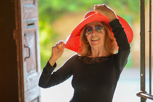 mature woman wearing a red hat glasses dark dress and dancing shoes strikes a confident sexy pose as she poses for a photograph.