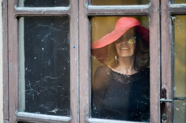 A woman with a hat on her head stands behind a window indoors while facing the camera,