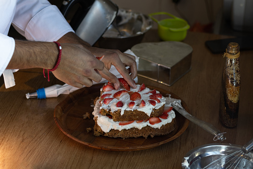 Horizontal photograph of an unrecognizable baker decorating his cake with strawberries and frosting, with his own hands, for a party