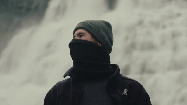 Handsome young man with winter hat and neck warmer looking at Icelandic waterfall