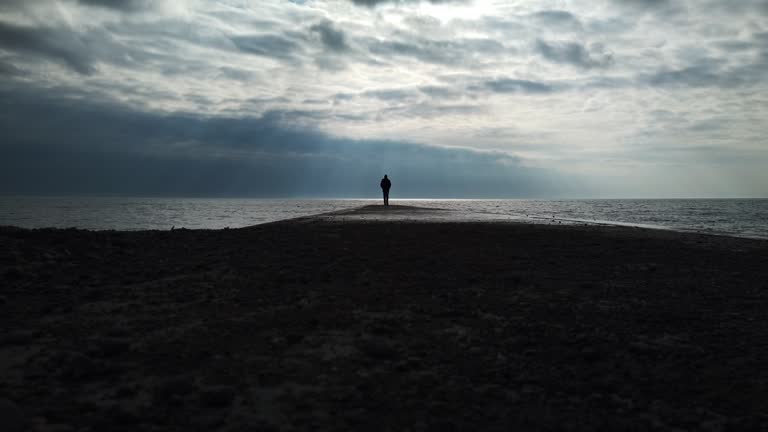 Man in a black coat walking on a concrete jetty to the infinite horizon of Lake Ontario