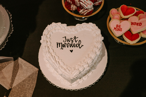Retro heart shaped cake with white frosting and glitter. “Just Married” written in black frosting across the middle of the cake.  Cake is placed on desert table with red and pink flowers, cookies, and flowers peddles.