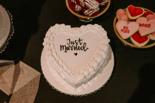 Retro “Just Married” heart shaped cake Retro heart shaped cake with white frosting and glitter. “Just Married” written in black frosting across the middle of the cake. Cake is placed on desert table with red and pink flowers, cookies, and flowers peddles. white wedding cake red roses stock pictures, royalty-free photos & images