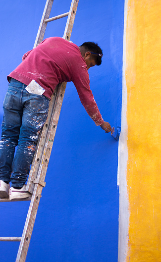 Oaxaca, Mexico: A man on a ladder painting a vibrant blue and yellow wall in downtown Oaxaca.