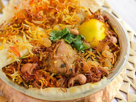 Hyderabadi chicken biryani with tomato, coriander, lemon, cashew nut and bread dum served in copper pot dish isolated on wooden table top view arabic spicy food