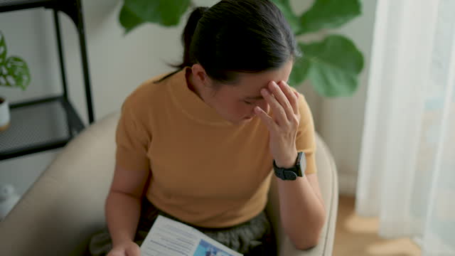 Asian woman sitting on armchair in living room at home stressed about bill payment problems.