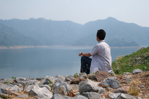Young Asian man looking away, sitting on rock over river, showing freedom on hill over reservoir and mountain view background, keep green to nature with human responsibility concept.