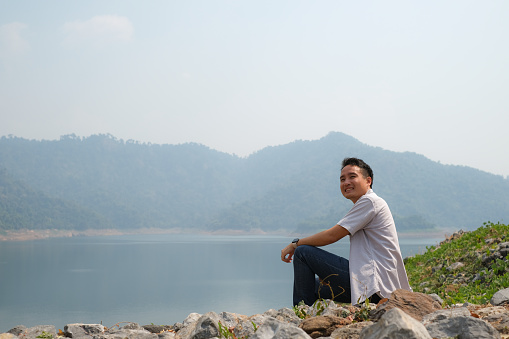 Young Asian man looking away, sitting on rock over river, showing freedom on hill over reservoir and mountain view background, keep green to nature with human responsibility concept.