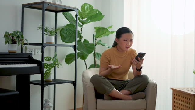 Asian woman feeling sadness reading or watching something at smartphone, sitting on armchair in living room at home.