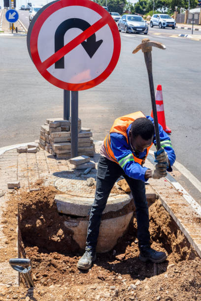 afroamerikanischer arbeiter mit einer spitzhacke, der einen graben in der autobahn aushebt, um kabel auf einer insel an einer kreuzung zu reparieren - construction safety mid adult men road construction stock-fotos und bilder