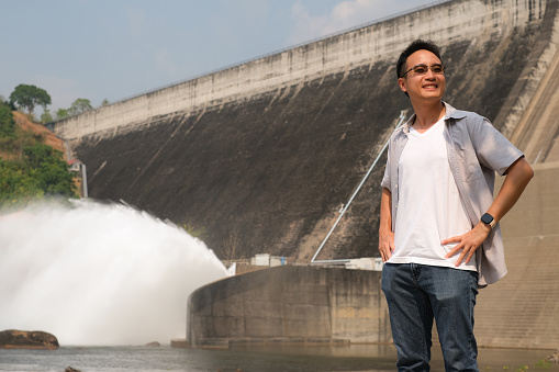 Young Asian handsome man standing looking away with feeling of why must to keep river clean and smile with water was spread out of dam to give power for human, people responsibility to nature concept.