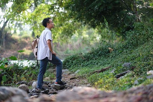 Young Asian man walking to the forest, looking up on walk to see the soundness of nature, nature surrounding him with forest, trees, water with little smile and feel freshness, people responsibility to nature concept.