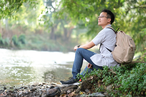 Young Asian man sitting on the rock by the river, looking up, see the soundness of nature, nature surrounding him with forest, trees, water with little smile and feel freshness, people responsibility to nature concept.