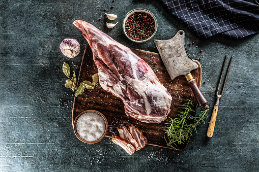 Raw lamb thigh surrounded by salt, pepper, garlic, bacon and rosemary on a vinateg surface.