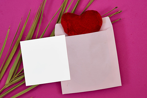 Wedding invitation card mockup and red envelope with red heart toy and palm leaf on pink background. Blank card mockup