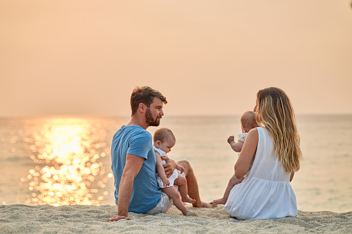 A family enjoying a tropical paradise beach during sunset. Mother and father are sitting with their twin girls and watching beautiful sunset.
