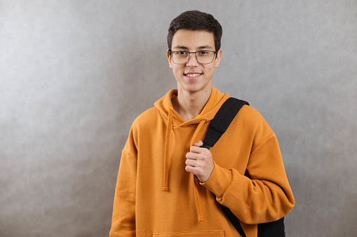 Portrait of a young student with a school bag. The teenager smiles and looks at the camera. A happy teenage boy on a grey background. Close up