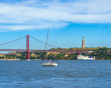 Sailboat and white ferry sailing along the Tajo River with the 25 de Abril steel suspension bridge in Lisbon and the religious monument Sacred Heart of Jesus of Almada in the background.