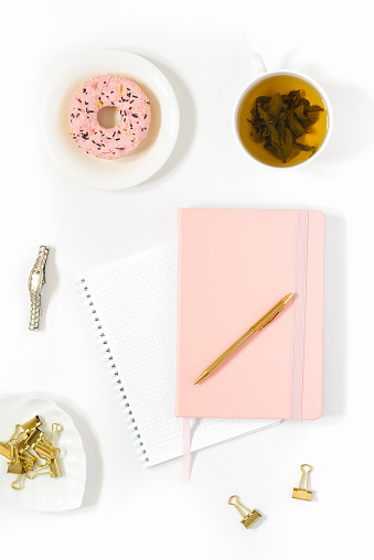 Stylish flat lay for a woman's fashion blogger. Workplace of a freelancer or office worker: Notepad and diary in a pink cover, a pen, a Cup of green tea, a pink donut. The trend concept, the vertical frame