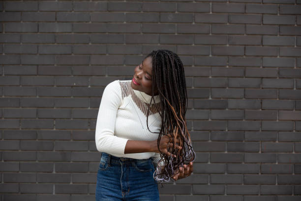 retrato da bela jovem mulher afro-americana mostrando cabelo preto longo trançado penteado enquanto se sente feliz e sorrindo na cidade ao ar livre - braided braids women long hair - fotografias e filmes do acervo