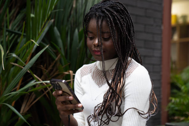 portrait of beautiful young african american woman showing long black hair braided hairstyle and using mobile phone while feeling happy and smiling in the city outdoor - braided braids women long hair imagens e fotografias de stock