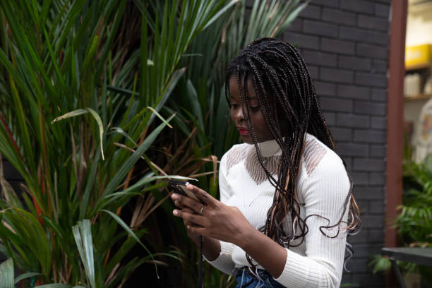 retrato da bela jovem mulher afro-americana mostrando cabelo preto longo trançado penteado e usando o telefone celular enquanto se sente feliz e sorrindo na cidade ao ar livre - braided braids women long hair - fotografias e filmes do acervo