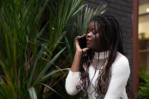 retrato da bela jovem mulher afro-americana mostrando cabelo preto longo trançado penteado e usando o telefone celular enquanto se sente feliz e sorrindo na cidade ao ar livre - braided braids women long hair - fotografias e filmes do acervo