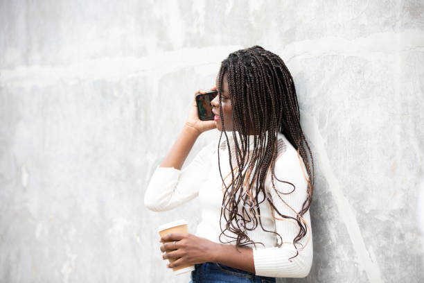 retrato da bela jovem mulher afro-americana mostrando cabelo preto longo trançado penteado e usando o telefone celular enquanto se sente feliz e sorrindo na cidade ao ar livre - braided braids women long hair - fotografias e filmes do acervo