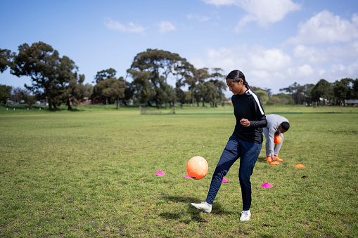 Girl soccer player practicing with ball with coach placing the training cones on ground in background on sports field