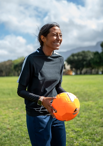 Happy young girl soccer player in sportswear holding ball on sports field looking away and smiling